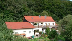 A two-story house with a red roof, surrounded by lush greenery and a forested hillside. - Quiet Holiday Home With Terrace (Korbach)