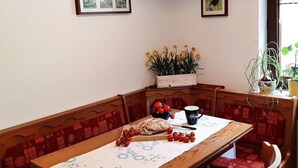 A dining area with a wooden table, red cushioned bench, and a white tablecloth. There are framed pictures on the wall, a potted plant, and a window with plants on the sill. - Quiet Holiday Home With Terrace (Korbach)
