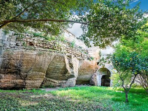 Une ancienne construction en pierre, dotée d’un portail en arc de cercle, et entourée d’une végétation luxuriante et d’arbres.