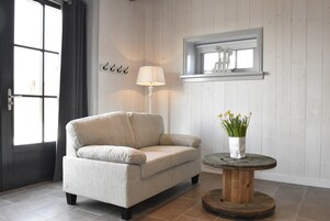 A room with a beige sofa, a round wooden table with a vase of yellow flowers, a lamp, and a window with a view.