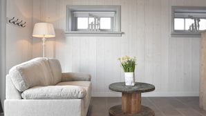 A room with a light-colored sofa, a round wooden table with a vase of yellow flowers, and two windows with blinds.