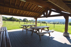 Een overdekte picknickplaats met houten banken en picknicktafels, gelegen op een geplaveid oppervlak met uitzicht op een grasveld en verre heuvels.