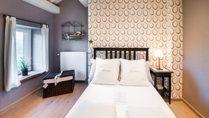 Een netjes opgemaakt bed met kussens met de tekst 'La Grande Randonnée', in een kamer met behang met een patroon, een nachtkastje met een lamp en een raam met gordijnen.