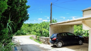 A black car parked under a concrete structure, a stone wall, and a road with greenery on both sides.