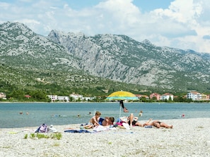 Ein Strand mit badenden Personen, einem gelben Sonnenschirm und Bergen im Hintergrund.