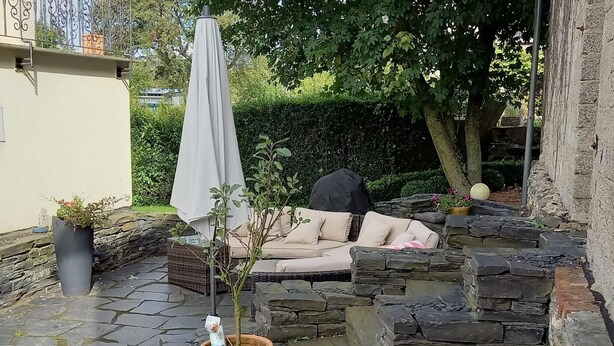A patio area with a stone wall, a beige sofa, and a potted plant. - Ferienhaus Paula in Haserich im Hunsrueck (Haserich)
