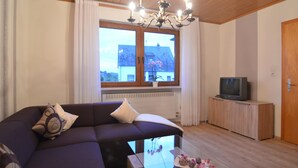 A living room with a sectional sofa, a glass coffee table, a television on a wooden cabinet, and a window with a view of a house. - Ferienhaus Paula in Haserich im Hunsrueck (Haserich)