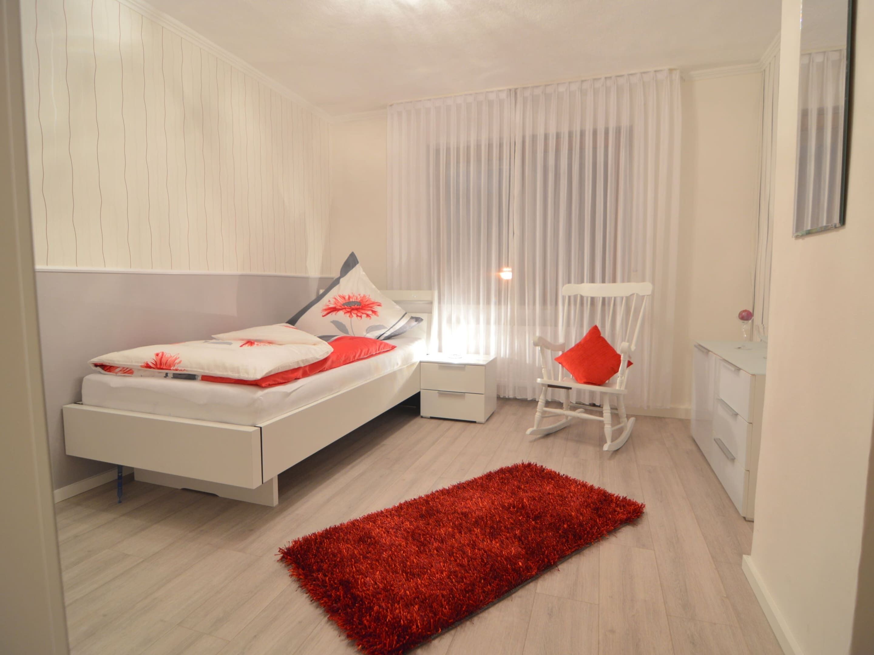 Un dormitorio moderno con cama, alfombra roja, una silla y una mesita de noche.
