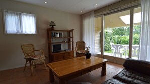 Sala de estar com mesa de madeira, duas cadeiras de vime, sofá de couro, aparador com televisão e luminária, e porta de vidro corrediça que dá para uma varanda com móveis de exterior.