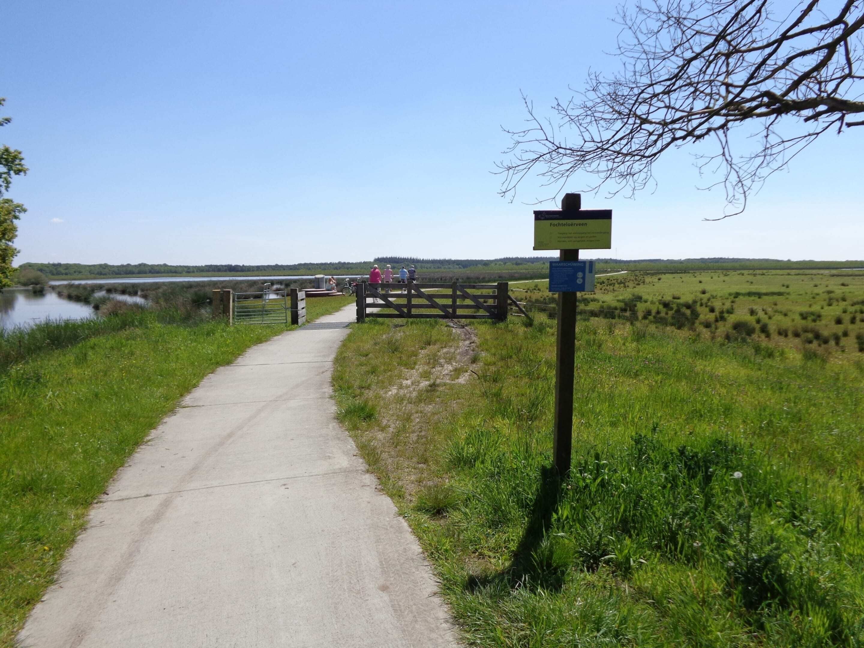 A concrete path leading to a wooden gate with a sign, surrounded by grassy fields and a body of water.
