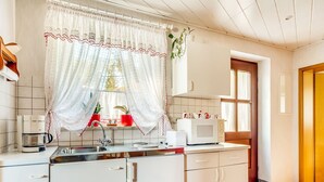 A modern kitchen with white cabinets, a stainless steel sink, a microwave, and a coffee maker. There are two red potted plants on the windowsill.