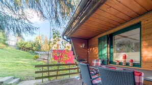 Une cabane en bois dotée d’un patio aménagé avec des meubles en osier, et un bâtiment rouge en arrière-plan.