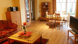 Una sala de estar con un sofá, una mesa de centro de vidrio, un televisor sobre un mueble bajo, una mesa de comedor con sillas, una estantería y una puerta que da a otra habitación.