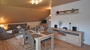 Ein modernes Wohnzimmer mit Holzboden, einer Couch, einem Esstisch mit Stühlen, einem Fernseher und einem Regal mit Dekoration.
