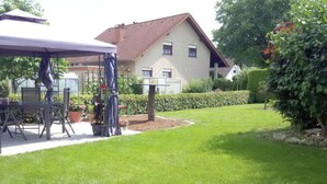 Ein gepflegter Garten mit einer Terrassenfläche, ein Backsteinhaus im Hintergrund und ein Heckenstreifen entlang der Grundstücksgrenze.