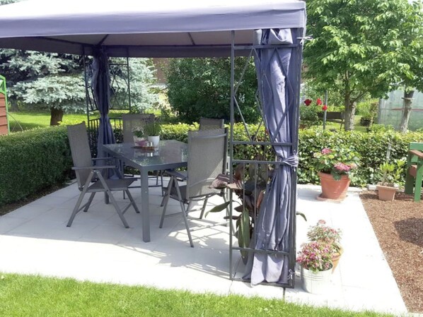 A covered patio area with a table and chairs, surrounded by potted plants and a garden.