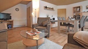 Ein modernes Wohnzimmer mit einem Glastisch, einer Couch, einem Esstisch mit Stühlen, einem Fernseher und einem Bücherregal.