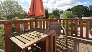 A wooden deck with a table and a wicker chair, an umbrella, and a view of a residential area with trees and houses.
