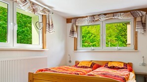 A bedroom with a wooden bed, two windows with curtains, a radiator, and a view of greenery outside. - Attractive Apartment in Rubeland in the Upper Harz (Oberharz am Brocken)