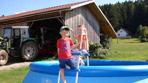 Dos niños en un flotador azul para piscina con una escalera, un tractor y un granero de madera al fondo.