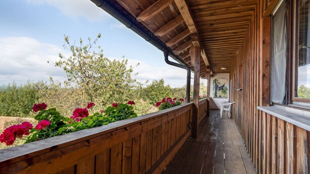 Una terraza de madera con barandilla, una mesa y sillas, con vista a un jardín con flores rojas y árboles.