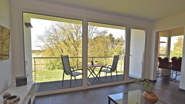 A modern living room with a glass table and chairs, large sliding glass doors leading to a balcony with a view of trees and a grassy area. - Das Kuenstlerhaus in Kammerstein (Kammerstein)