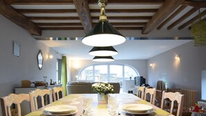 A dining area with a long table set for a meal, wooden chairs, a chandelier, and a view of the outdoors through large windows.
