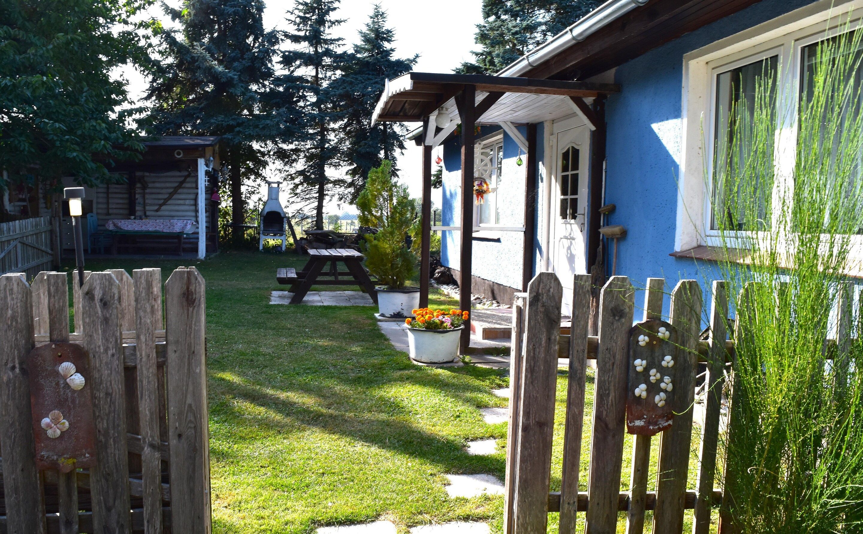 Una valla de madera con un portón, un patio cubierto de césped, una parrilla para barbacoa y una casa con fachada azul.