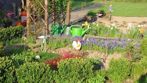 Ein Garten mit verschiedenen Pflanzen, einem Gießkanne und einer Hahn-Figur.