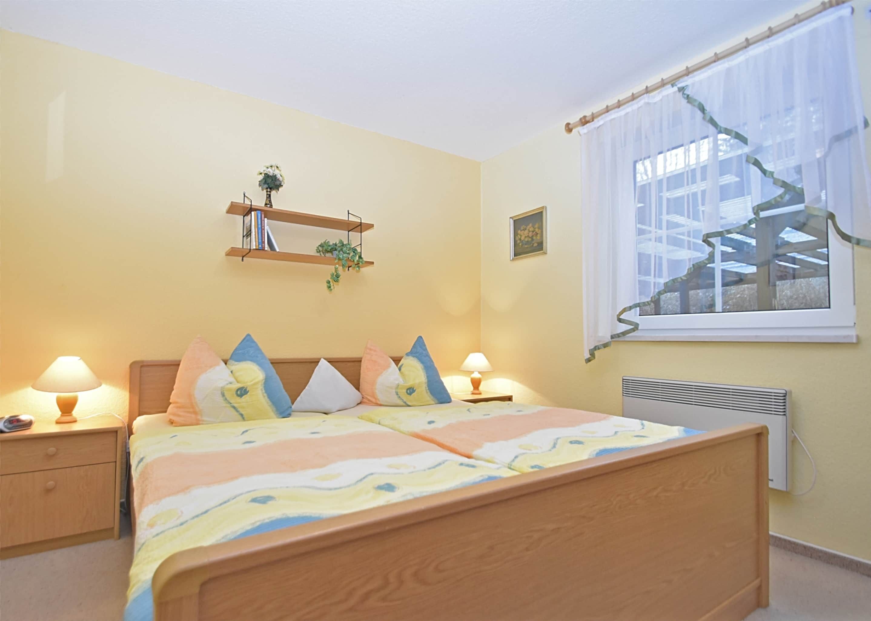 A bedroom with a bed, bedside lamps, a window with curtains, a radiator, a shelf with books and a plant, and a picture on the wall.