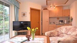 A living room with a sofa, a coffee table with a book and a vase of flowers, a television, and a kitchen with wooden cabinets and a window.