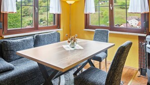 A dining area with a table and chairs, two windows with curtains, a lamp, and a view of greenery outside. - Quiet Home Near Vessertal (Schleusegrund)