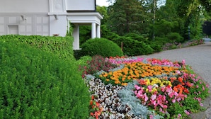 En vit byggnad med en täckt veranda, omgiven av frodig grönska och en färgstark blomsterträdgård.