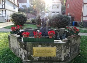 A statue of a woman in a garden with a plaque, surrounded by flower beds and houses in the background. - Wohnung in Bad Grund (Bad Grund)