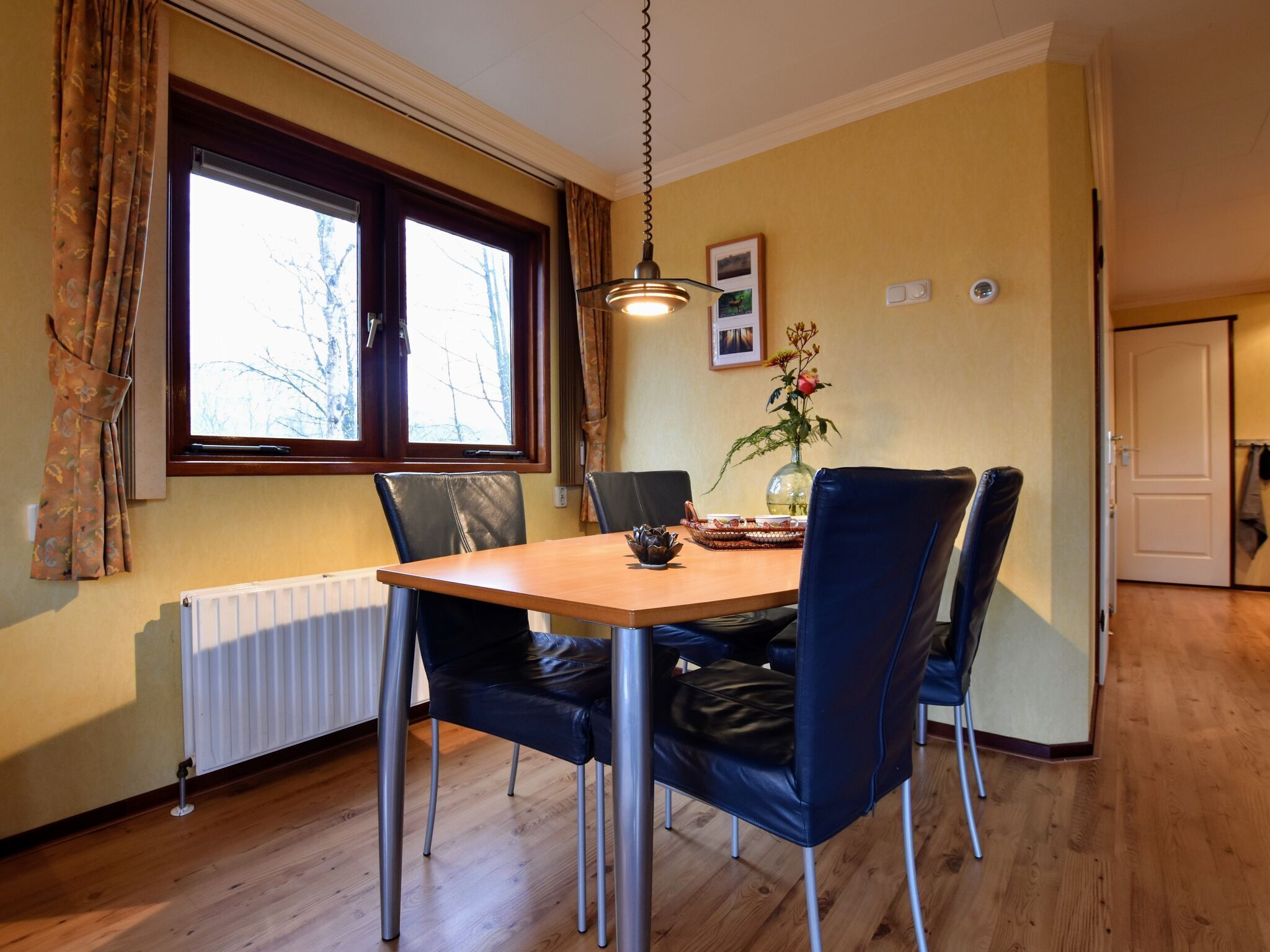 Un coin repas avec une table en bois, des chaises en cuir noir, un vase de fleurs et une fenêtre avec des rideaux.