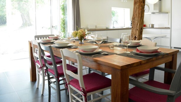 Een eetruimte met een houten tafel voor zes personen, omringd door witte stoelen met rode kussens. Een grote boomstam staat in het midden van de kamer, en er is uitzicht op een tuin door de open deur.