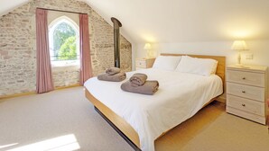 A bedroom with a sloped ceiling, stone walls, a large window with curtains, a bed with white linens, two bedside lamps, and a wooden nightstand.