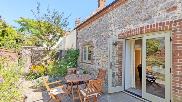 A stone building with a patio area featuring a wooden table and chairs, a glass door, and a garden with various plants and flowers.