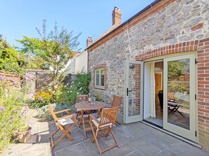 A stone building with a patio area featuring a wooden table and chairs, a glass door, and a garden with various plants and flowers. - Hearnesbrook Barn , Emsworth - an annexe that sleeps 4 guests in 2 bedrooms (Emsworth)