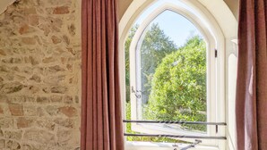 A room with a stone wall, a window with a metal railing, and a view of greenery outside.