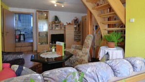 Uma sala de estar com sofá, mesa de centro e estante de livros. Escada leva a um andar superior.