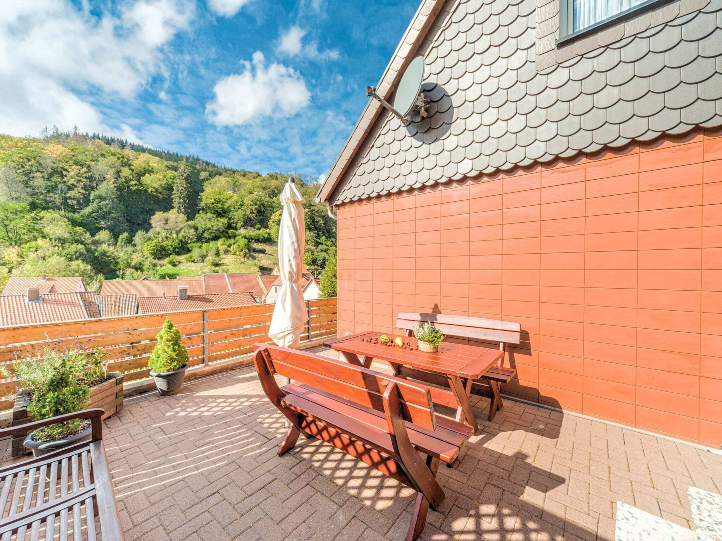 A patio with wooden benches and a table, surrounded by potted plants and a view of a forested hillside.