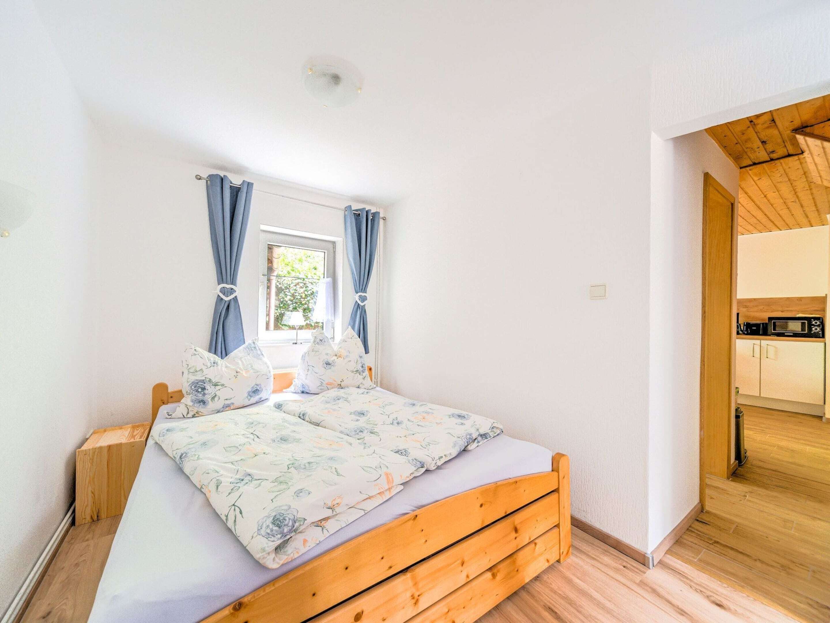 A bedroom with a wooden bed, a window with curtains, and a view of a kitchen.