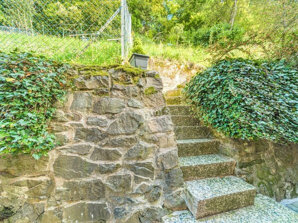 En stentrappe fører op ad en stenbelagt sti, omgivet af frodig vegetation og et metalhegn.