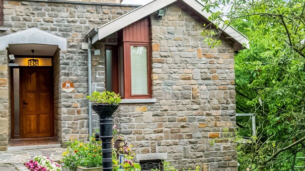 A stone building with a wooden door, a window with a red frame, and a planter with green plants. - A Small Studio of a Nature Stone House (Aywaille)