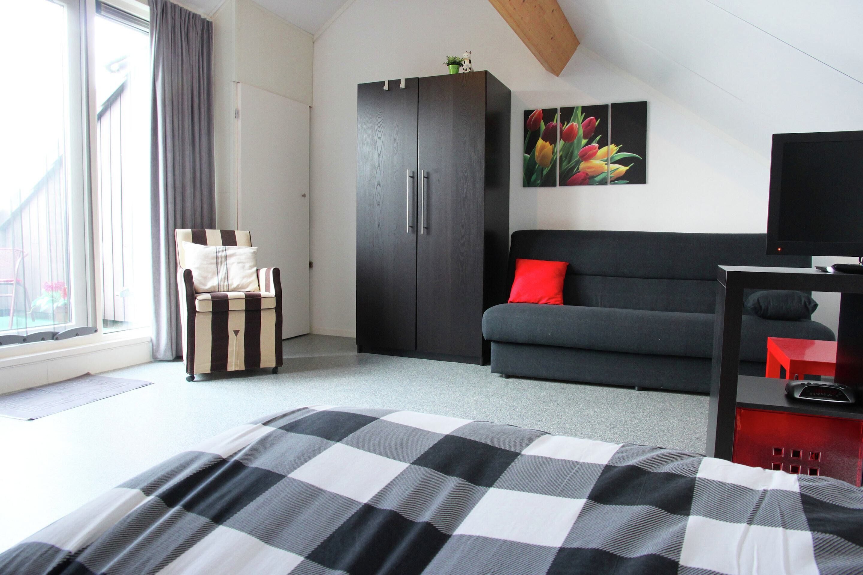 Une chambre à coucher avec un lit, un canapé gris, une armoire sombre, un coussin rouge, une télévision et une fenêtre avec des rideaux.