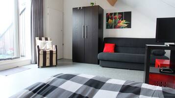 Une chambre à coucher avec un lit, un canapé gris, une armoire sombre, un coussin rouge, une télévision et une fenêtre avec des rideaux.