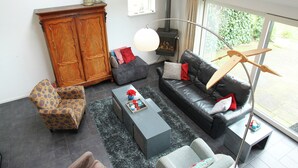 Ein Wohnzimmer mit Sofa, Sessel und Schrank. Ein Kamin, eine Lampe und ein Fenster mit Blick ins Grüne sind ebenfalls vorhanden.