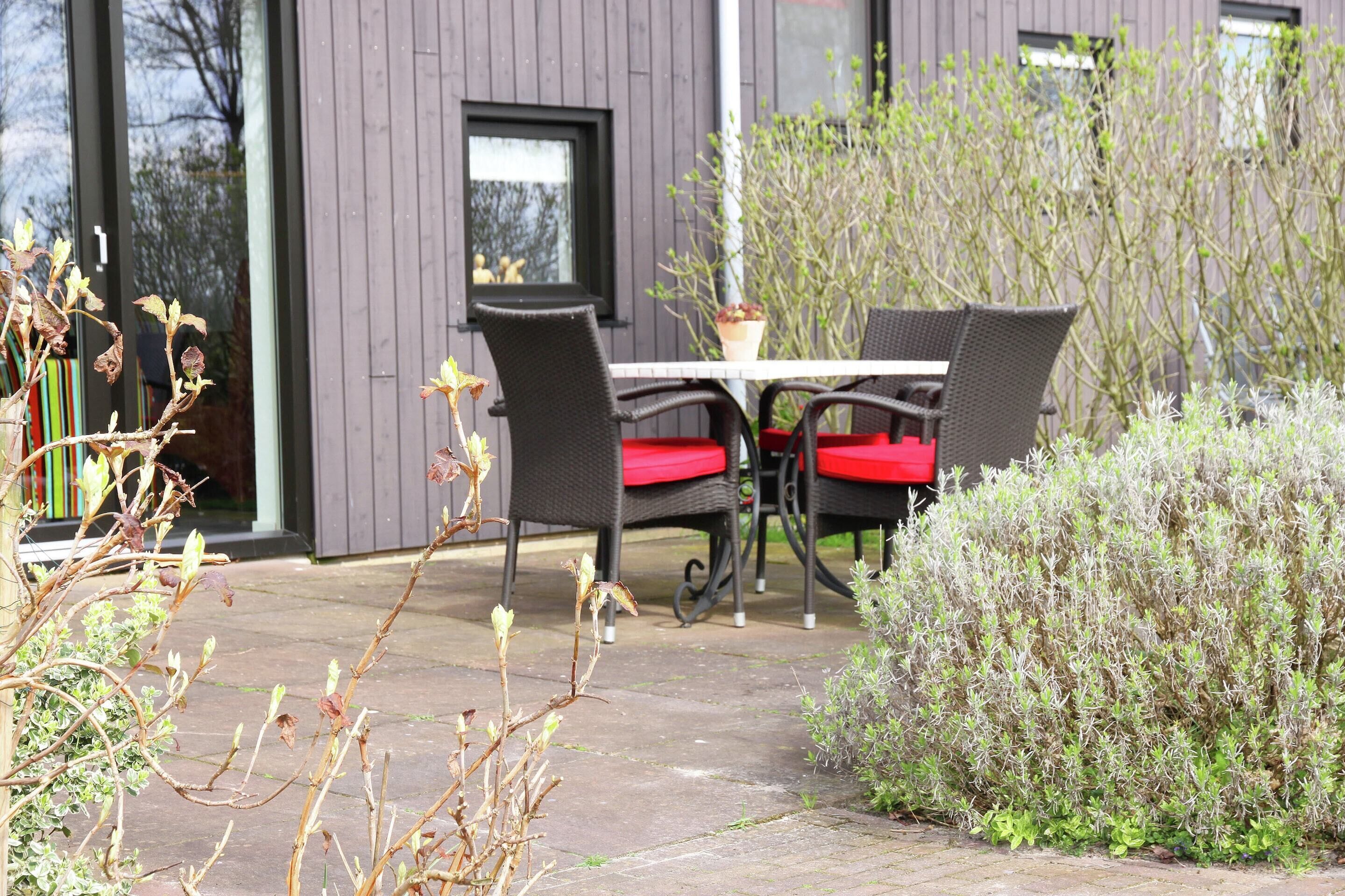 Une terrasse extérieure avec une table et des chaises, entourée de plantes et avec un bâtiment moderne en arrière-plan.