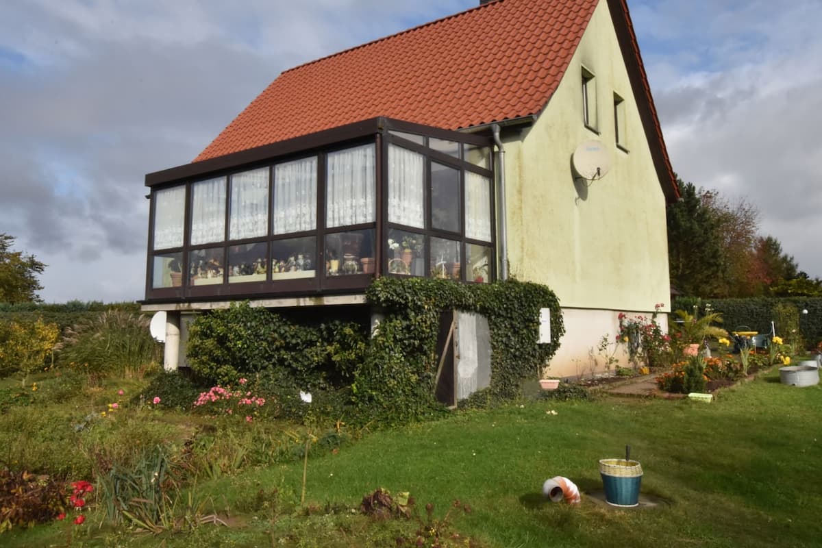 Ein traditionelles Haus mit rotem Ziegeldach, großen Fenstern und einem Garten mit verschiedenen Pflanzen und Blumen.